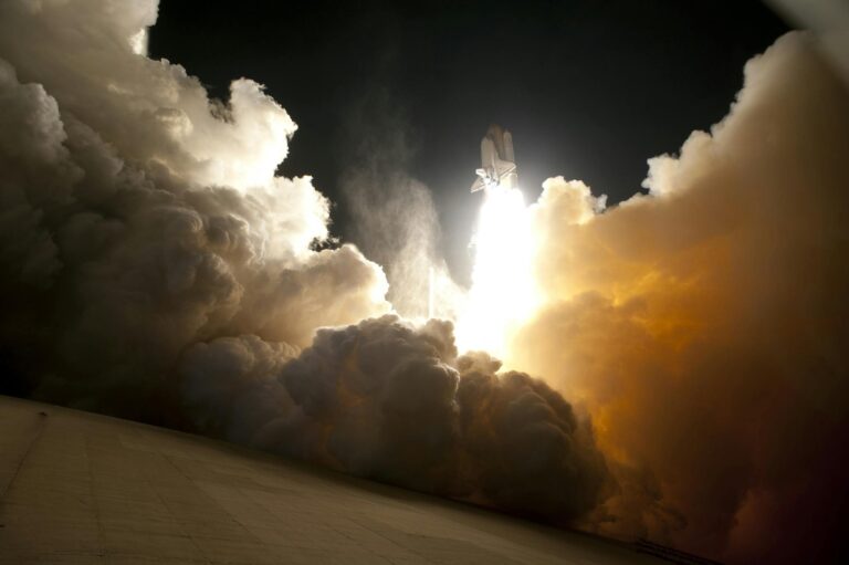 A dramatic image of a space shuttle launching into the night sky, surrounded by billowing smoke and intense light.